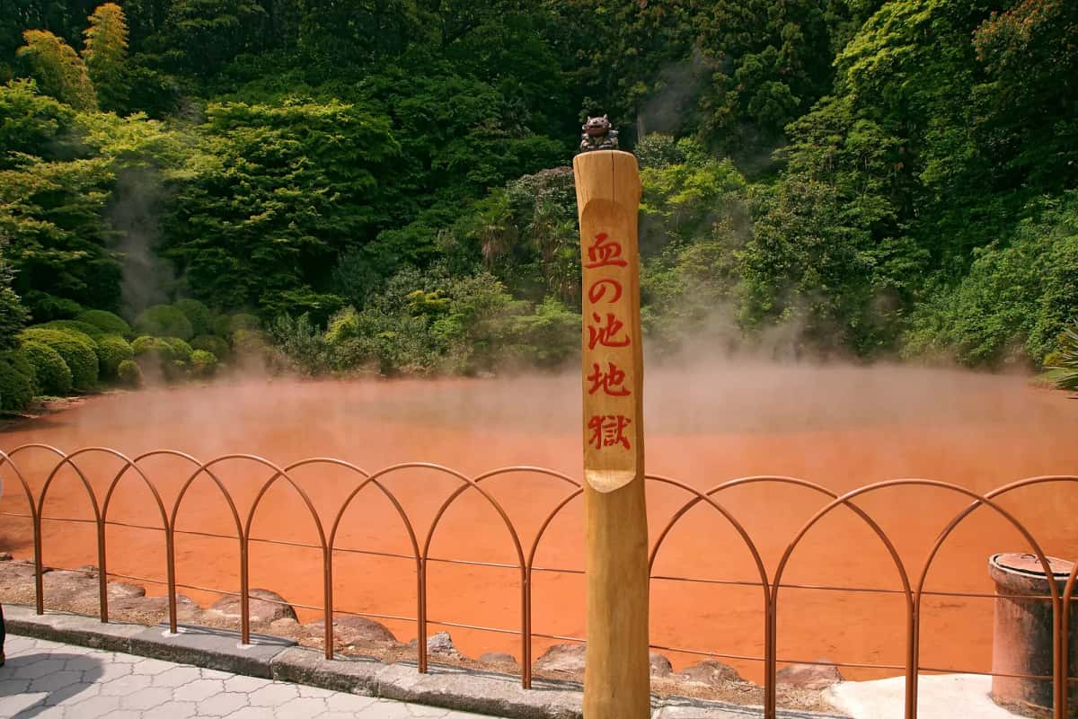 L'étang rouge fumant de Chinoike Jigoku derrière une barrière en arceaux et un poteau en bois gravé de son nom, à Beppu