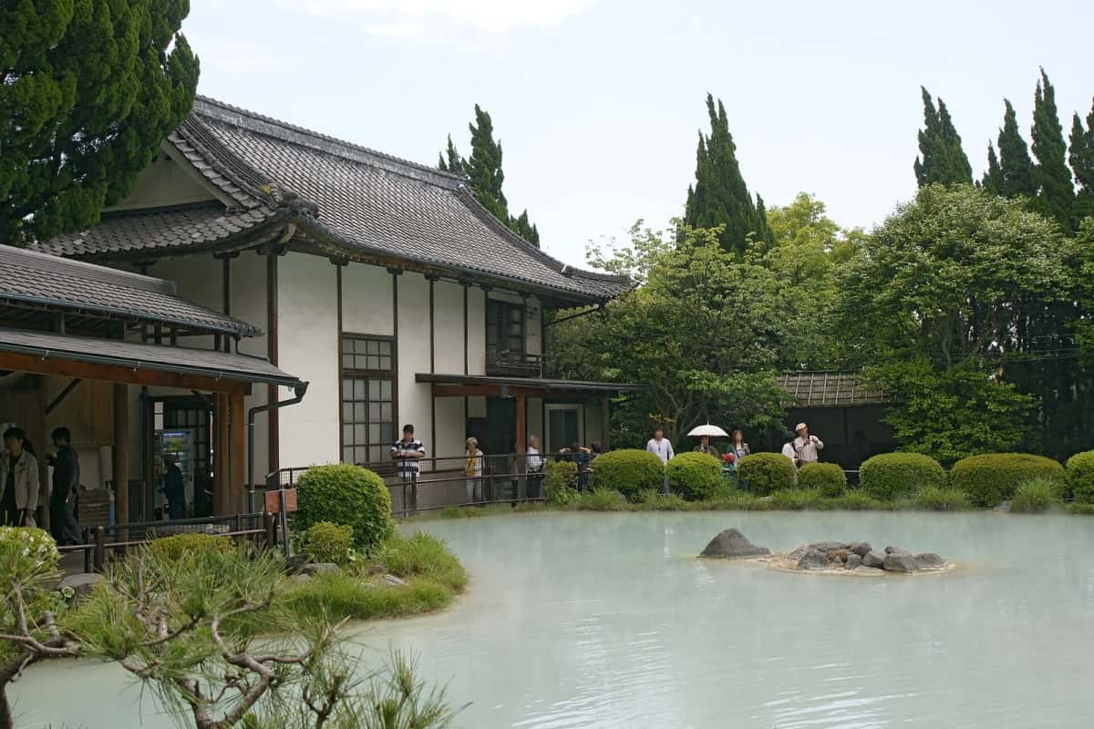 Le bassin d'eau blanc laiteux de Shiraike Jigoku devant un bâtiment traditionnel et un jardin de pins, à Beppu