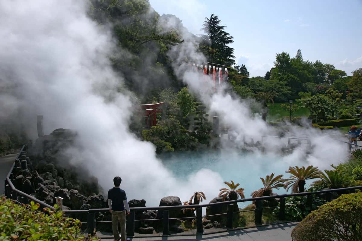 Le bassin bleu turquoise d'Umi Jigoku sous des nuages de vapeur, avec un torii rouge et un visiteur au bord, à Beppu