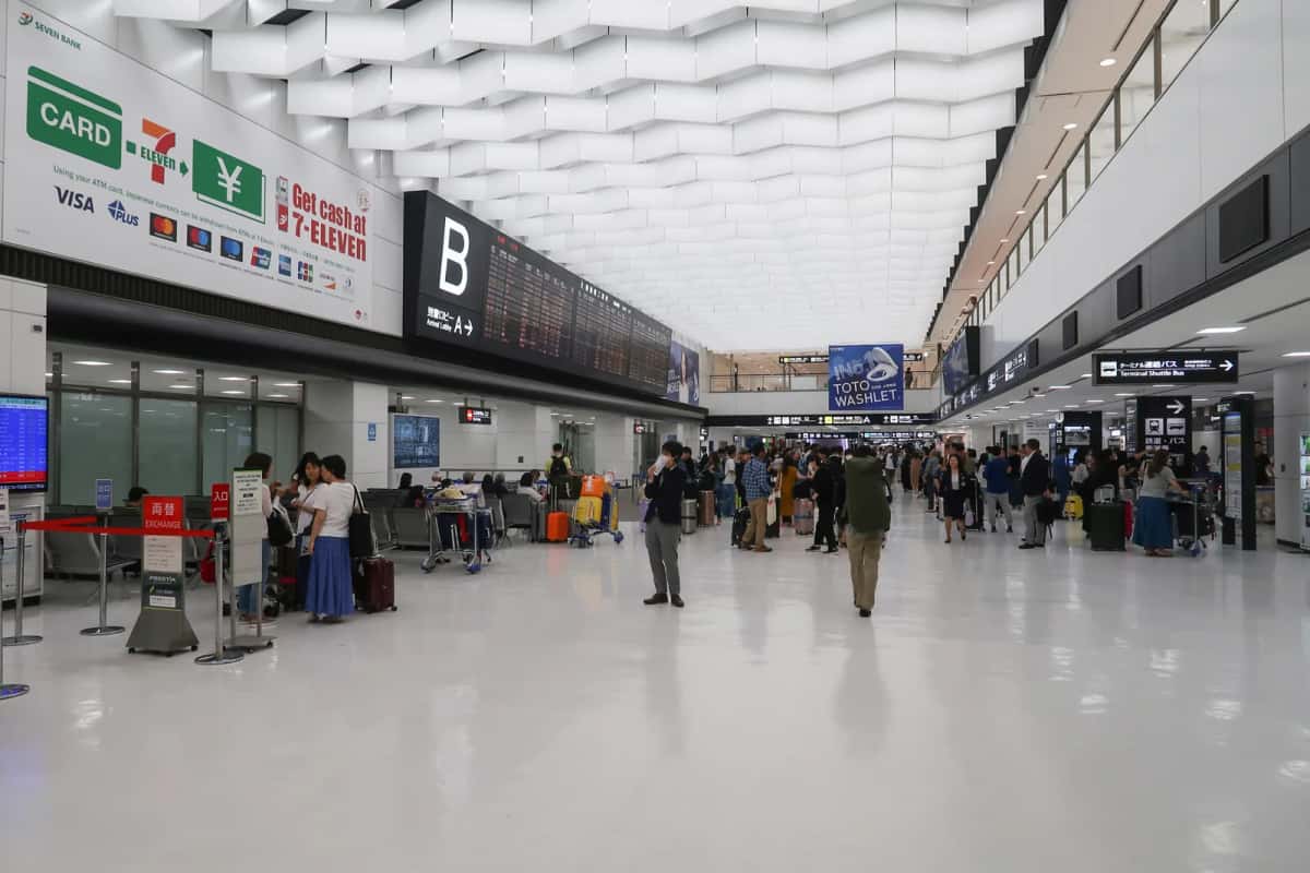 Hall des arrivées du terminal 2 de l'aéroport de Narita, avec comptoirs, panneaux en japonais et en anglais et voyageurs avec valises