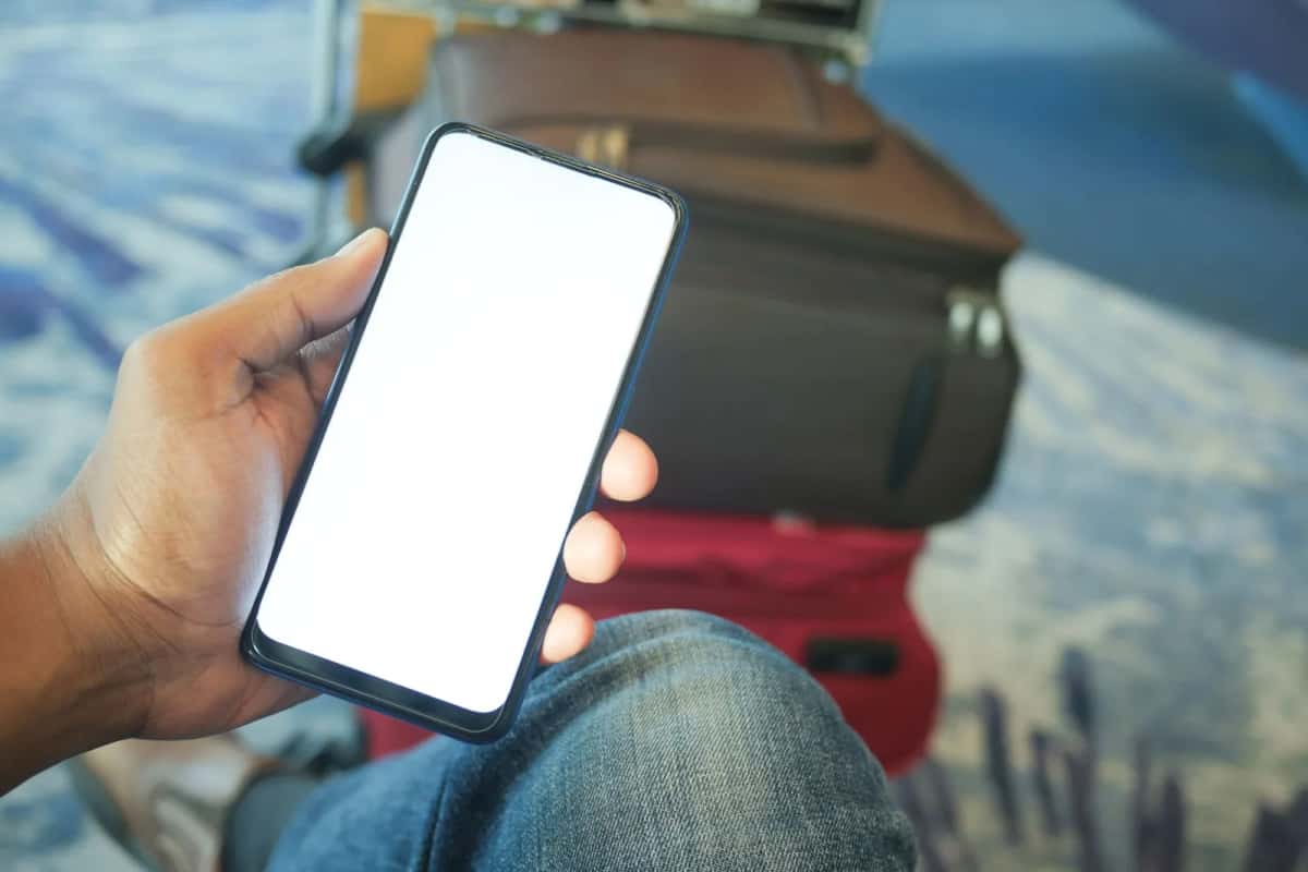 Mains tenant un smartphone à l'écran allumé, une valise en arrière-plan, pendant la préparation d'un voyage