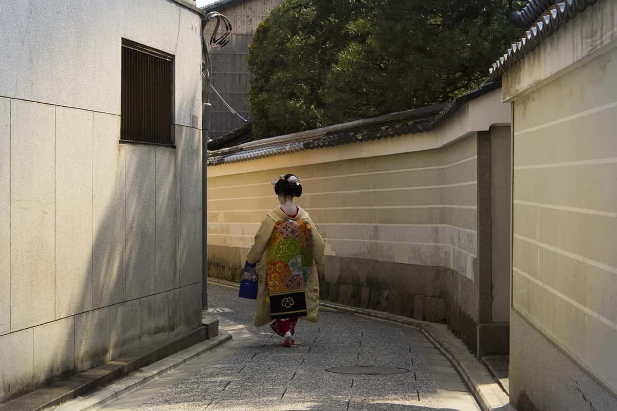 Une maiko marchant dans une ruelle du quartier Kamishichiken à Kyoto, le long d'un mur blanc