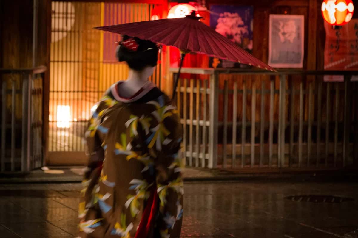 Une maiko en kimono sous une ombrelle rouge, de dos, dans la rue Hanamikoji-dori à Gion, le soir