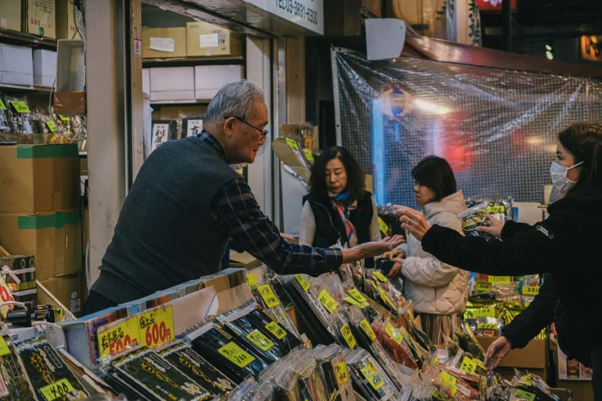 Commerçant âgé et clientes à un étal de marché au Japon, étiquettes de prix en yens