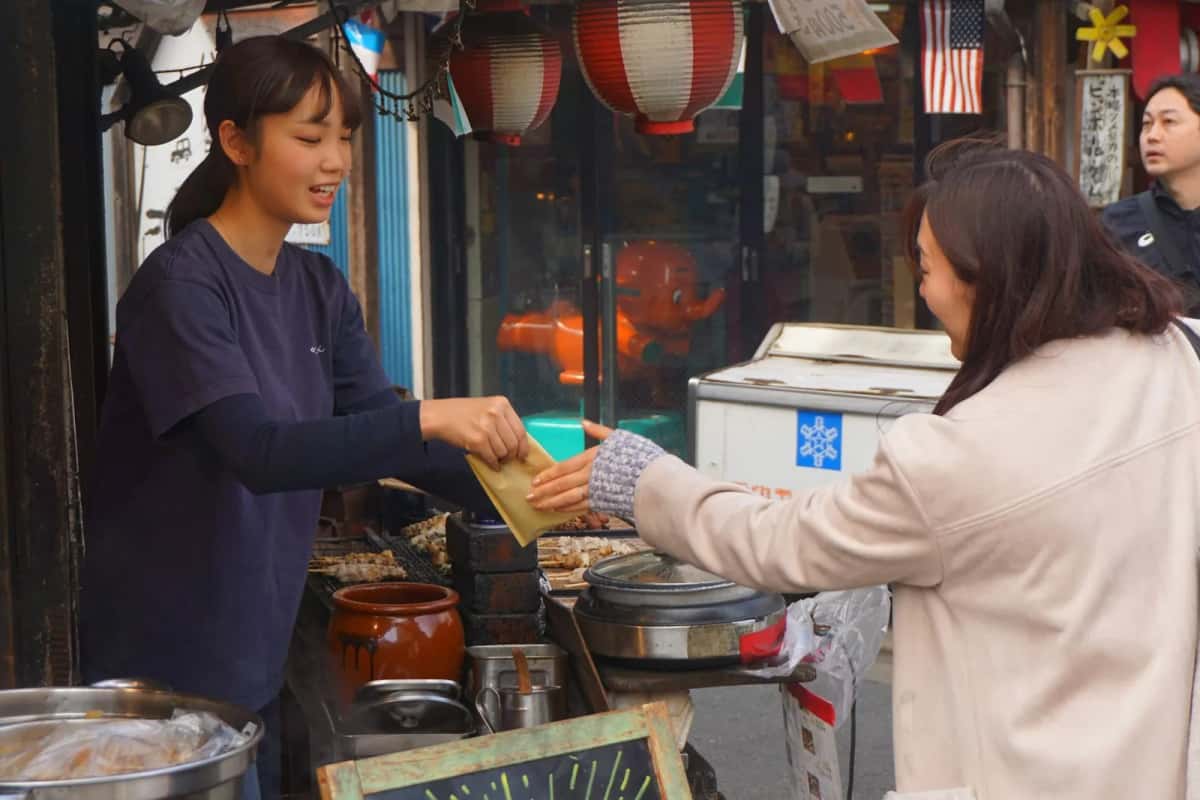 Vendeuse souriante tendant un paquet à une cliente à l'étal d'une rue commerçante de Tokyo
