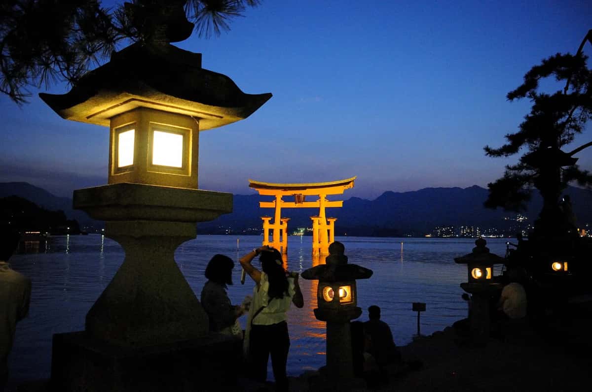 Le grand torii d'Itsukushima illuminé de nuit derrière une lanterne de pierre allumée, Miyajima