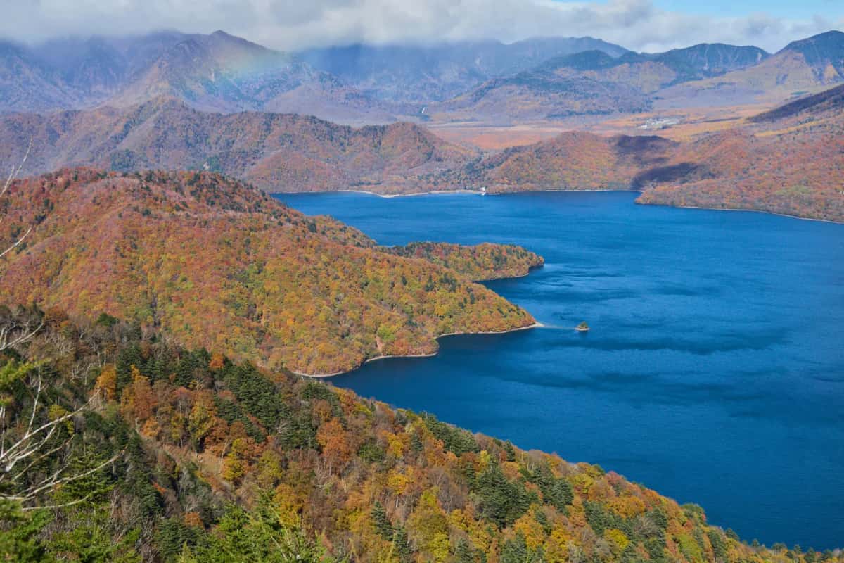 Le lac Chūzenji bleu profond entre des montagnes couvertes de forêts rousses et dorées, vu d'un col en automne
