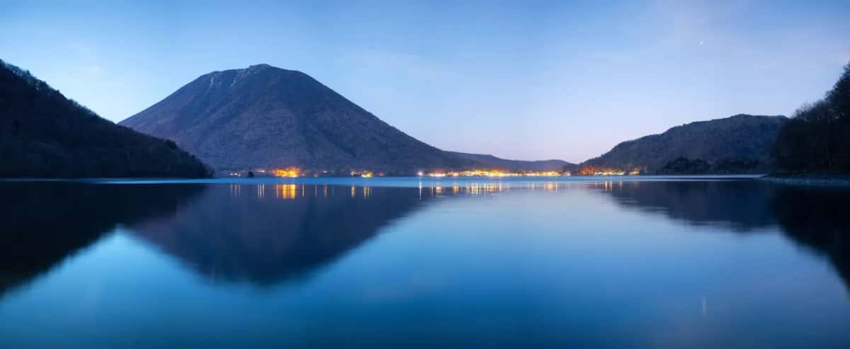Le lac Chūzenji à la tombée de la nuit, le mont Nantai reflété dans l'eau et les lumières du village au bord