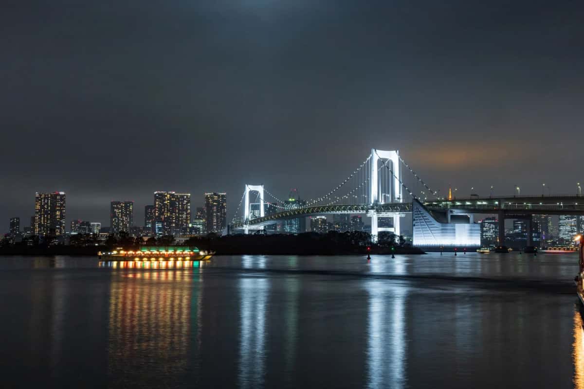 Le Rainbow Bridge illuminé en blanc la nuit, qui se reflète dans la baie de Tokyo devant les tours de Shibaura