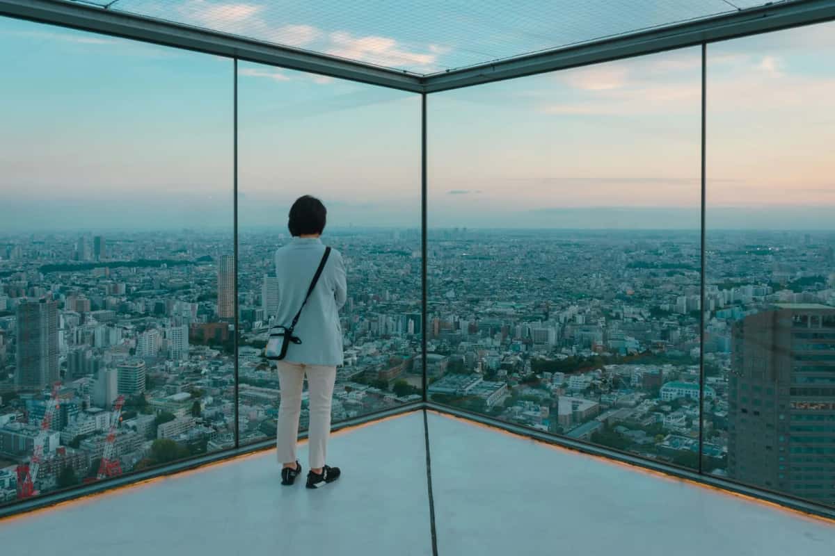 Visiteuse dans l'angle vitré du Sky Edge de Shibuya Sky, face à Tokyo au crépuscule