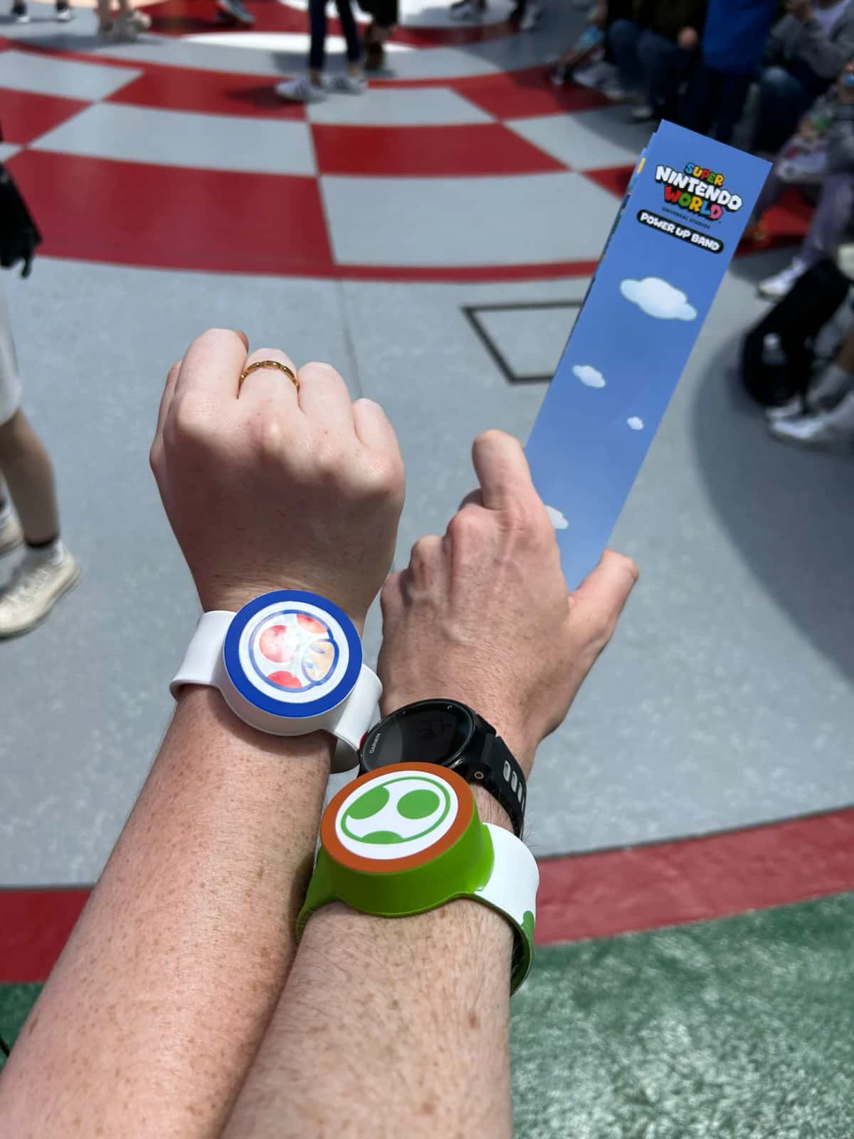 Deux Power-Up Band aux couleurs de Mario et de Yoshi portés au poignet, sur le sol en damier de Super Nintendo World